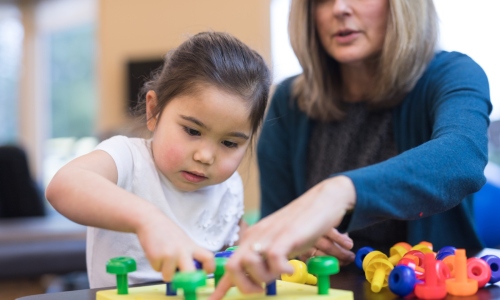An occupational therapist works with a kindergarten-age ethnic girl on her coordination skills. She is showing her a fun exercise  to practice putting pegs into a plastic board.