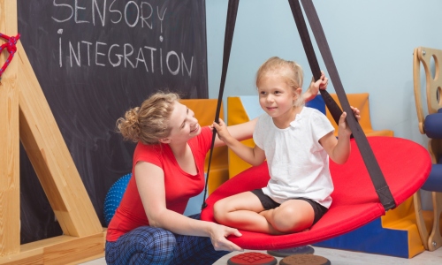 Shot of a smiling girl enjoying a sensory therapy on a swing while her physiotherapist assisting her