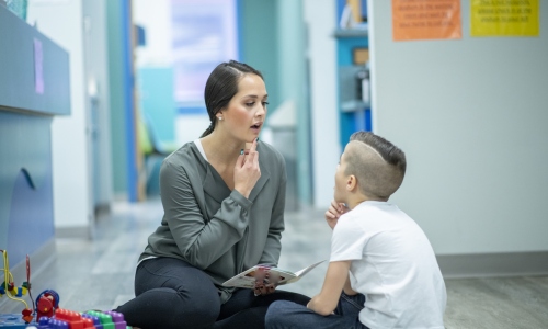 A young female Speech Therapist sits on the floor with her hand on her chin as she helps her young patient with his speech. She is dressed in semi-casual clothing and working with the young boy using a story book that is open in her hand.  The boy is dressed casually and seated cross-legged on the floor as he looks up at the therapist and copies her words with his hand on his own chin.