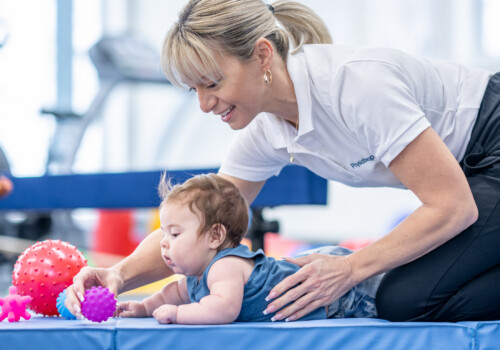 A female physical therapist works with an infant during a therapy session. She has the baby boy laying on a mat on his tummy as she helps him work on his core strength and gross motor skills with a ball. infant doing tummy time in physical therapy