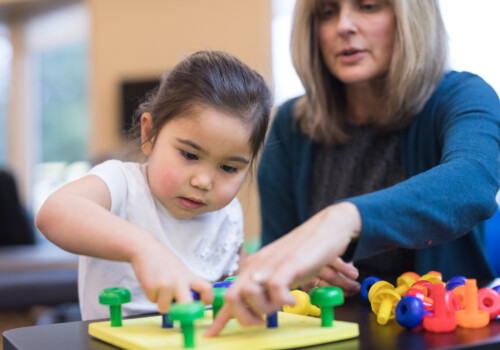 An occupational therapist works with a kindergarten-age ethnic girl on her coordination skills. She is showing her a fun exercise to practice putting pegs into a plastic board. pediatric occupational therapy