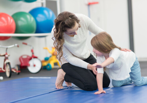 A kindergarten-age girl does exercises with her physical therapist at a clinic. She is doing a balancing exercise on the mat while the female therapist helps support her. There is a rack of medicine balls and play equipment in the. background, including a pair of tricycles.