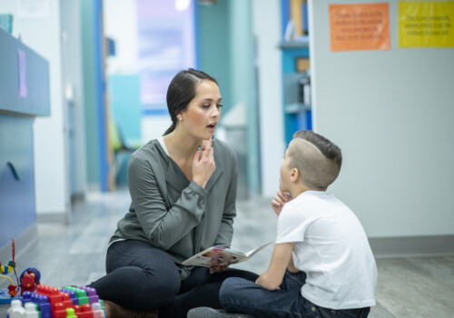 A young female Speech Therapist sits on the floor with her hand on her chin as she helps her young patient with his speech. She is dressed in semi-casual clothing and working with the young boy using a story book that is open in her hand. The boy is dressed casually and seated cross-legged on the floor as he looks up at the therapist and copies her words with his hand on his own chin. speech therapy with toys