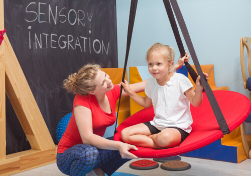 Shot of a smiling girl enjoying a sensory therapy on a swing while her physiotherapist assisting her sensory swing in occupational therapy